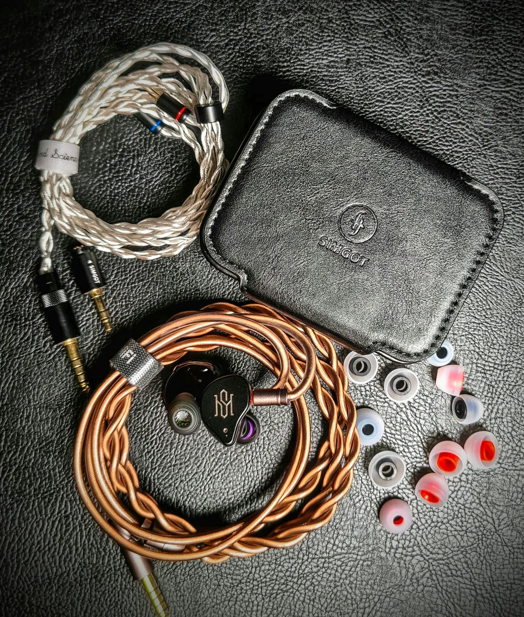 An organized top-down overview of the Simgot SuperMix 5 accessories and unboxing package arranged on a black textured leather surface. The shot displays a braided silver "Sound Science" cable, a rectangular black faux-leather protective case embossed with the Simgot logo, the main braided copper cable attached to the IEM shell, and multiple pairs of Type 07 silicone eartips with distinct red and black inner stems.