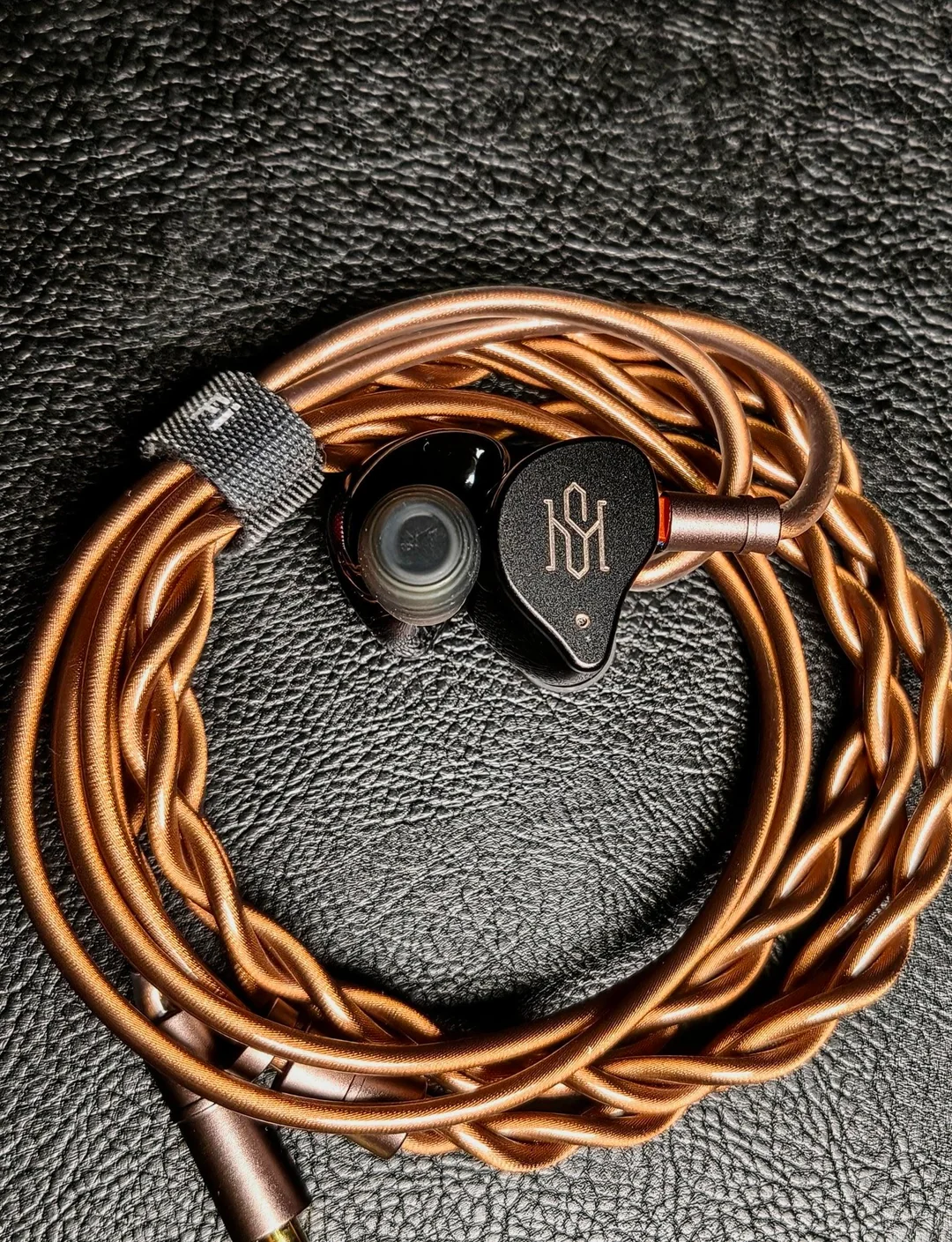 Uma foto em close-up de um único fone de ouvido intra-auricular Simgot SuperMix 5 (IEM), com faceplate de metal preto texturizado apresentando um logotipo geométrico dourado estilizado. O corpo do fone é de resina preta brilhante/transparente, permitindo ver os drivers internos, e está equipado com uma ponteira de silicone cinza. Ele repousa sobre um cabo entrançado de cobre, com um fecho de velcro preto com a inscrição "Fi" visível, tudo sobre uma superfície de couro preto. Esta imagem mostra o design e os detalhes do driver BCD analisado no review.
