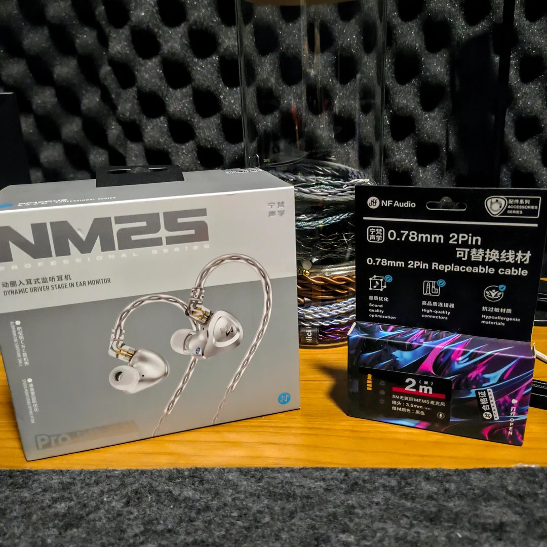 A front-facing shot of the left and right NF ACOUS NM25 IEM shells resting on a dark gray textured surface. The silver aluminum faceplates display a stylized engraved logo, with gold QDC-style 2-pin connectors pointing upward. Color-coded rings (green and purple) are visible near the nozzles. The official NM25 product box is blurred in the background, showcasing the compact and sturdy design.