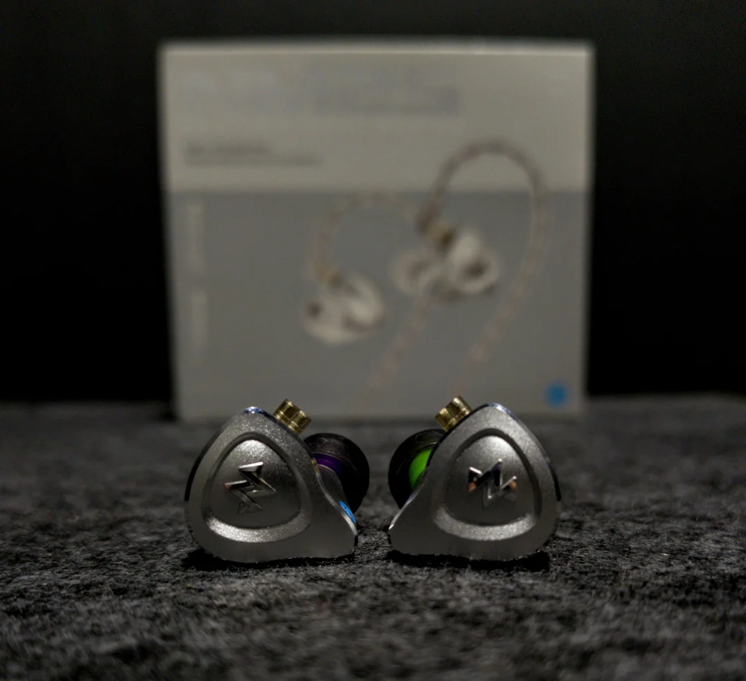 A front-facing shot of the left and right NF ACOUS NM25 IEM shells resting on a dark gray textured surface. The silver aluminum faceplates display a stylized engraved logo, with gold QDC-style 2-pin connectors pointing upward. Color-coded rings (green and purple) are visible near the nozzles. The official NM25 product box is blurred in the background, showcasing the compact and sturdy design.