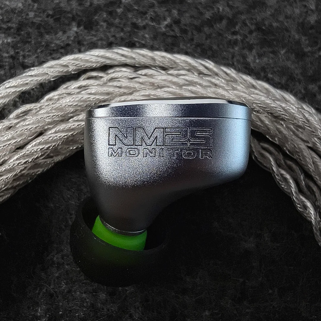 A close-up macro shot of the NF ACOUS NM25 in-ear monitor shell. The matte silver aviation aluminum housing features "NM25 MONITOR" engraved precisely on its side. It is fitted with a neon green MS42 silicone eartip and rests against a blurred braided silver audio cable on a dark textured background, highlighting the 5-axis CNC machined build quality.