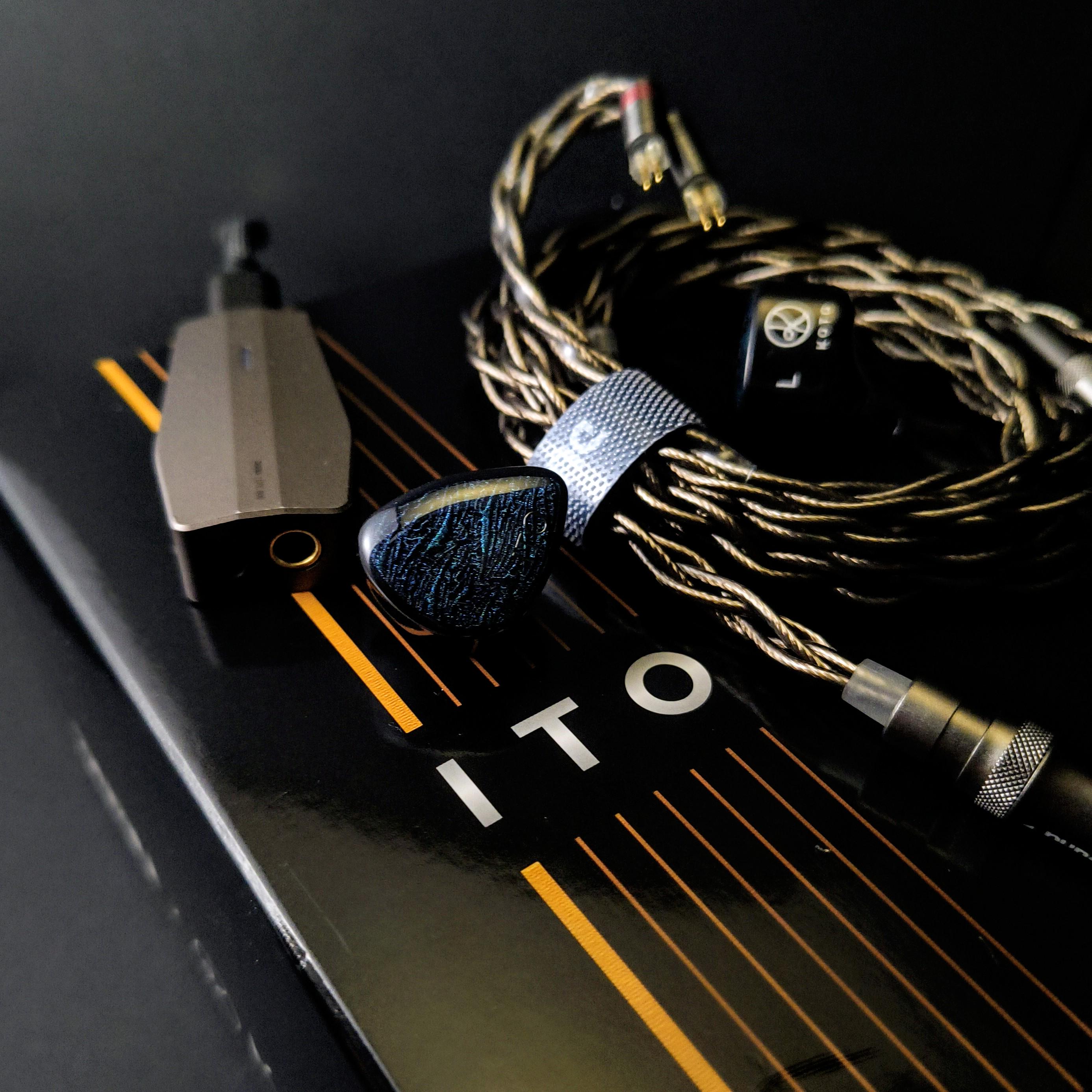 A close-up shot of the DUNU x KOTO ITO in-ear monitor resting on its black and gold retail box. The IEM features a stunning dark resin shell with a subtle bluish-green wavy faceplate pattern. Alongside it is the premium braided "LEO" copper cable and a grey portable DAC/AMP dongle, highlighting the ideal audiophile setup for this bass-heavy set.