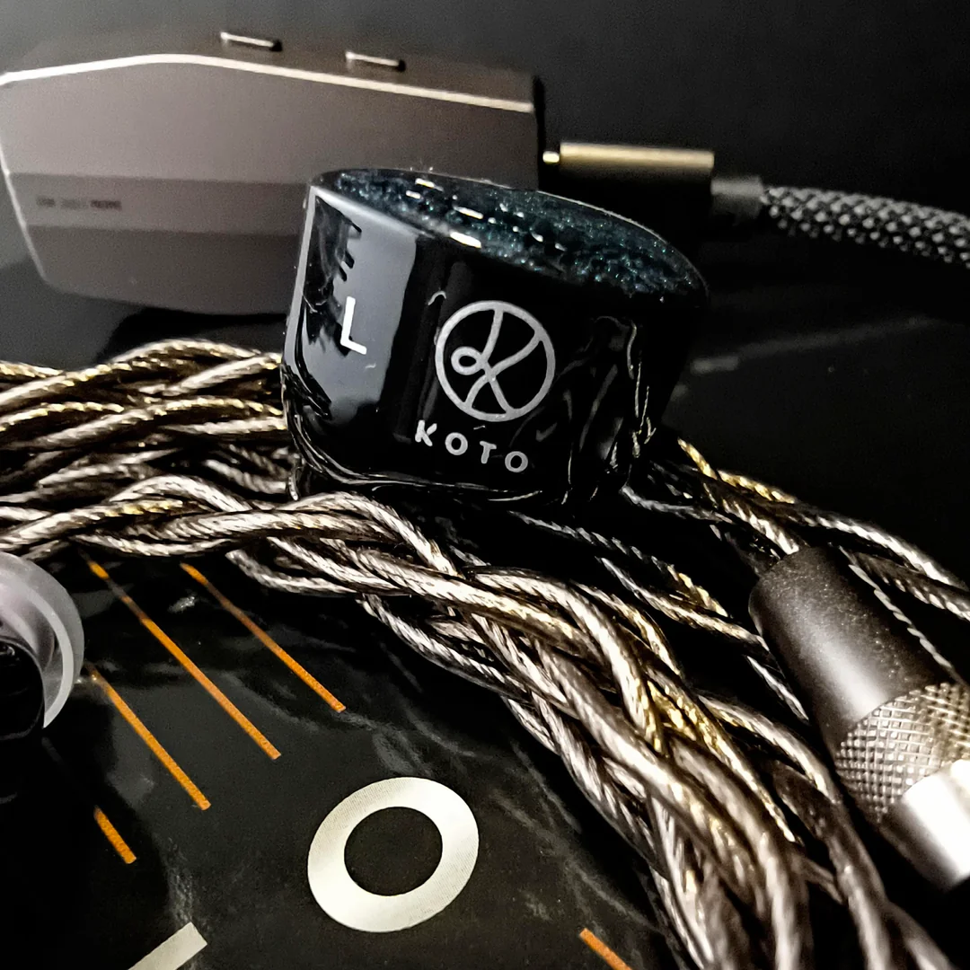 A macro side-profile shot of the DUNU x KOTO ITO in-ear monitor. The glossy black resin shell prominently displays the white stylized KOTO logo and a Left "L" indicator. It rests on the premium braided "LEO" copper cable, with the black and gold ITO packaging and a blurred portable DAC/AMP in the background.