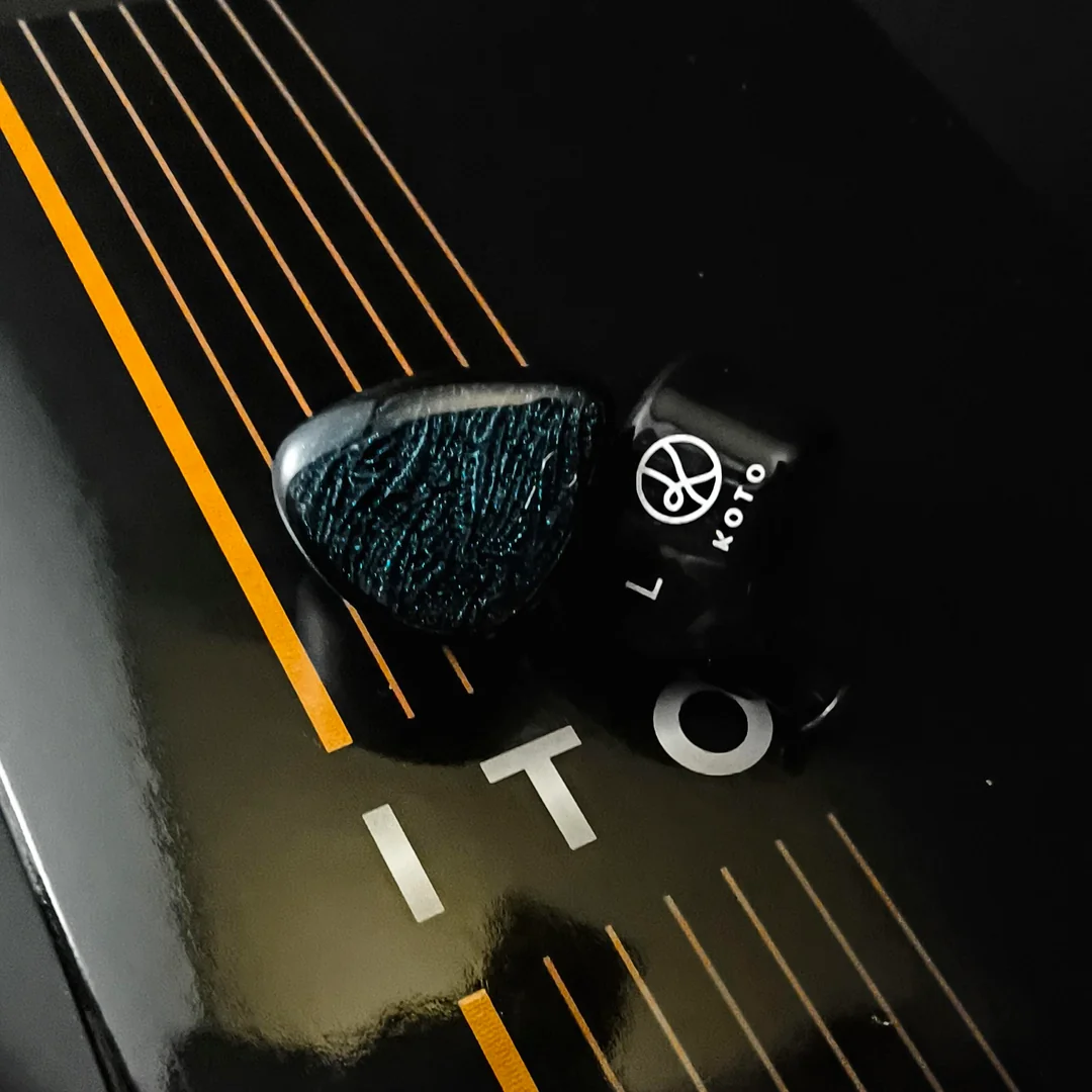 A close-up shot of two DUNU x KOTO ITO IEM shells resting directly on their official black and gold packaging. The left shell reveals the faceplate's subtle bluish-green wavy pattern under the light, while the right shell is positioned to show the white KOTO logo and "L" marker on its glossy black resin body.