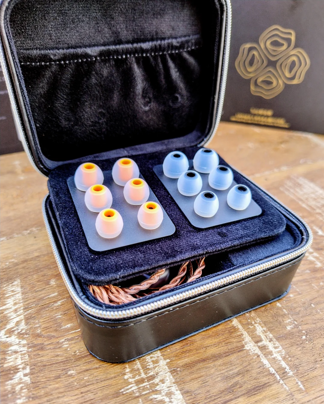 A close-up view of the open FAAEAL Tulip premium black leather carrying case. Inside the velvet-lined case, a display card elegantly holds six pairs of silicone eartips with orange and black inner stems. The braided copper audio cable is neatly tucked into the storage compartment below.