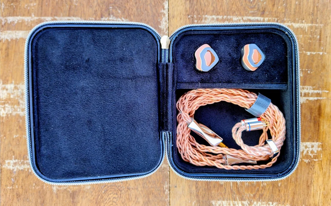 The fully open FAAEAL Tulip storage case resting on a textured wooden surface. The top section securely displays the two rose-gold and grey petal-shaped IEM shells, while the bottom section neatly contains the coiled "Hibiscus" copper cable and its polished metal hardware.