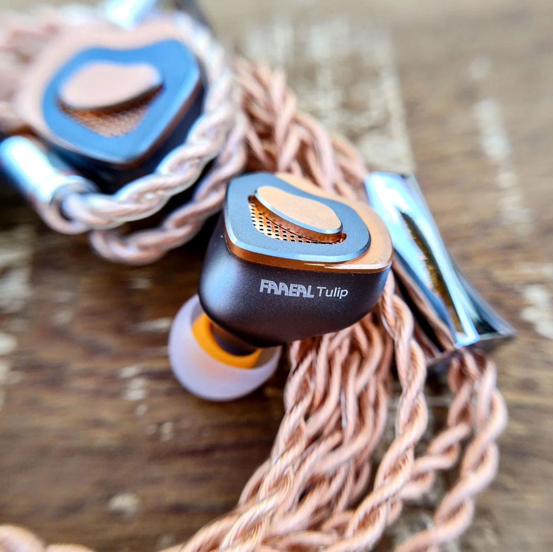 A macro shot of the FAAEAL Tulip in-ear monitor shell resting on its braided rose-copper audio cable. The dark grey CNC-machined aluminum body features the text "FAAEAL Tulip" cleanly printed on the side and is fitted with a translucent silicone eartip with an orange stem.