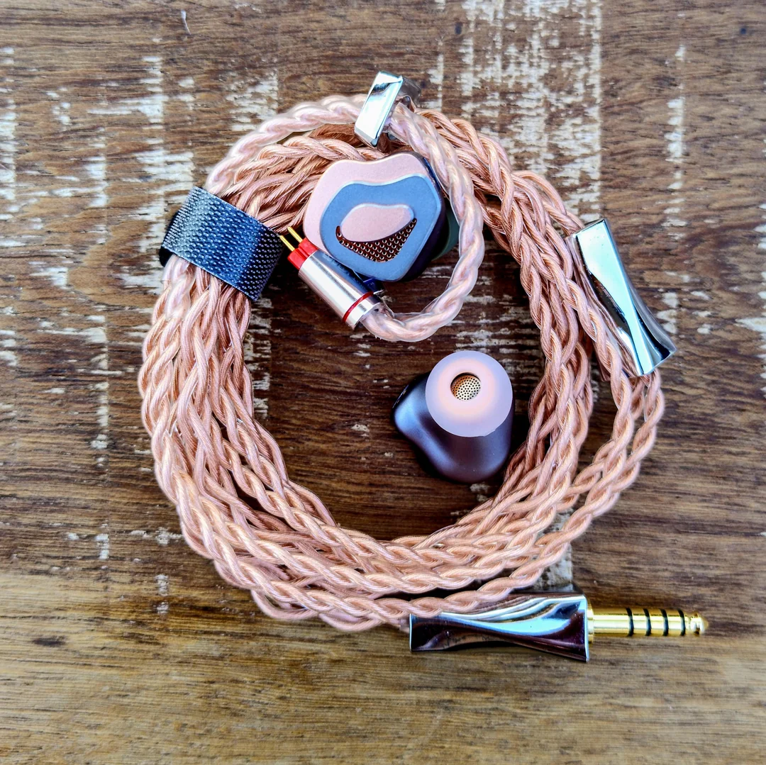 An aesthetically pleasing arrangement of the FAAEAL Tulip audio setup. The high-quality rose-copper "Hibiscus" cable is coiled circularly on a wooden surface, showcasing its shiny metal splitter and 4.4mm balanced plug. The two IEM shells rest inside the coil, displaying both the faceplate design and the inner side with an attached eartip.