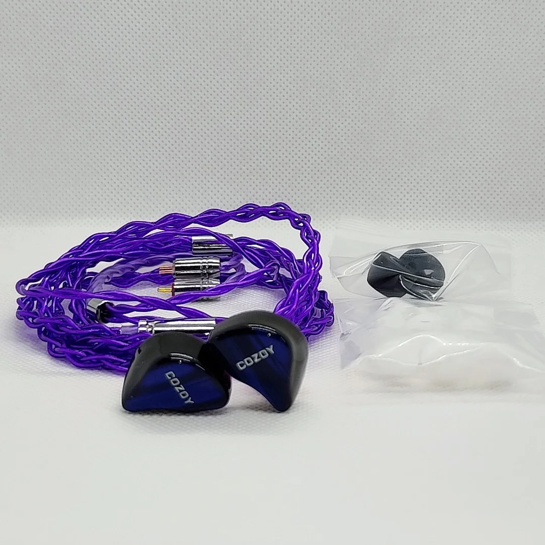 The Cozoy D1 IEMs and their vibrant purple braided audio cable resting on a white background. They are displayed next to two small, sealed clear plastic bags containing the included stock silicone eartips.