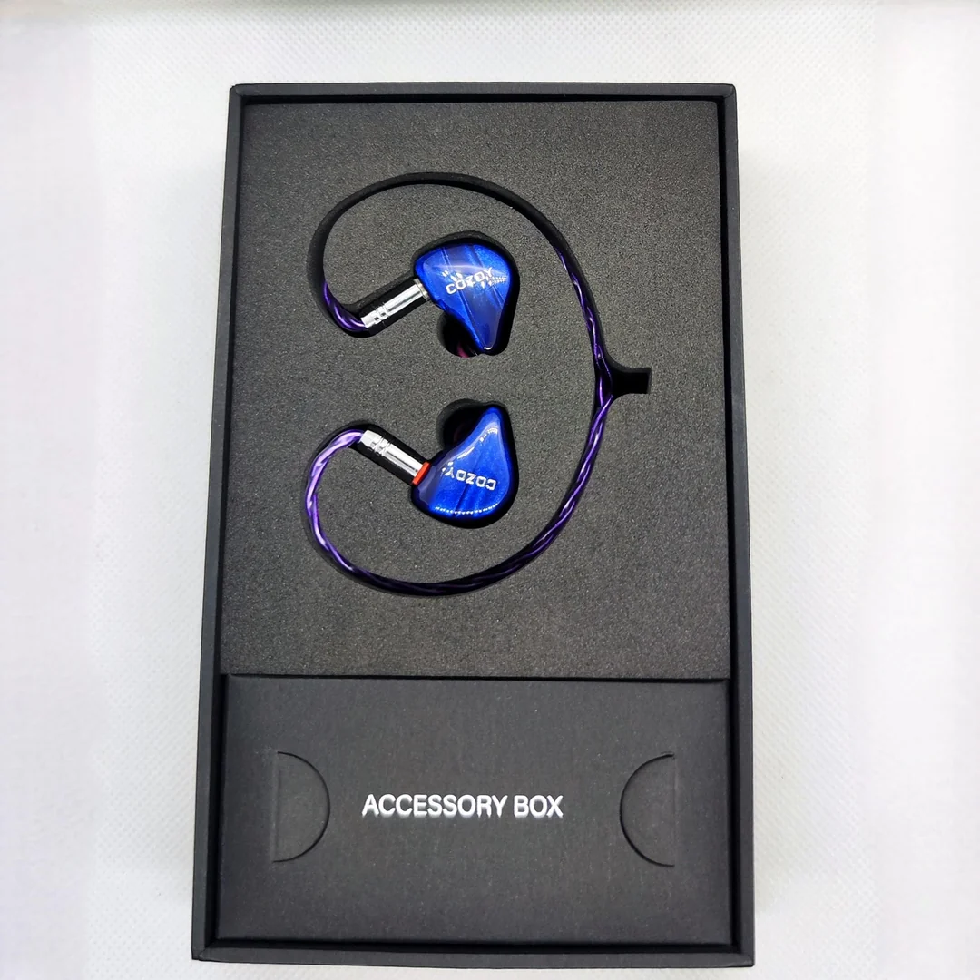 A top-down view of the Cozoy D1 in-ear monitors neatly displayed inside their black foam retail packaging. The glossy blue resin shells with the silver Cozoy logo are attached to a vibrant purple braided cable. The lower section of the foam insert features a compartment clearly labeled "ACCESSORY BOX".