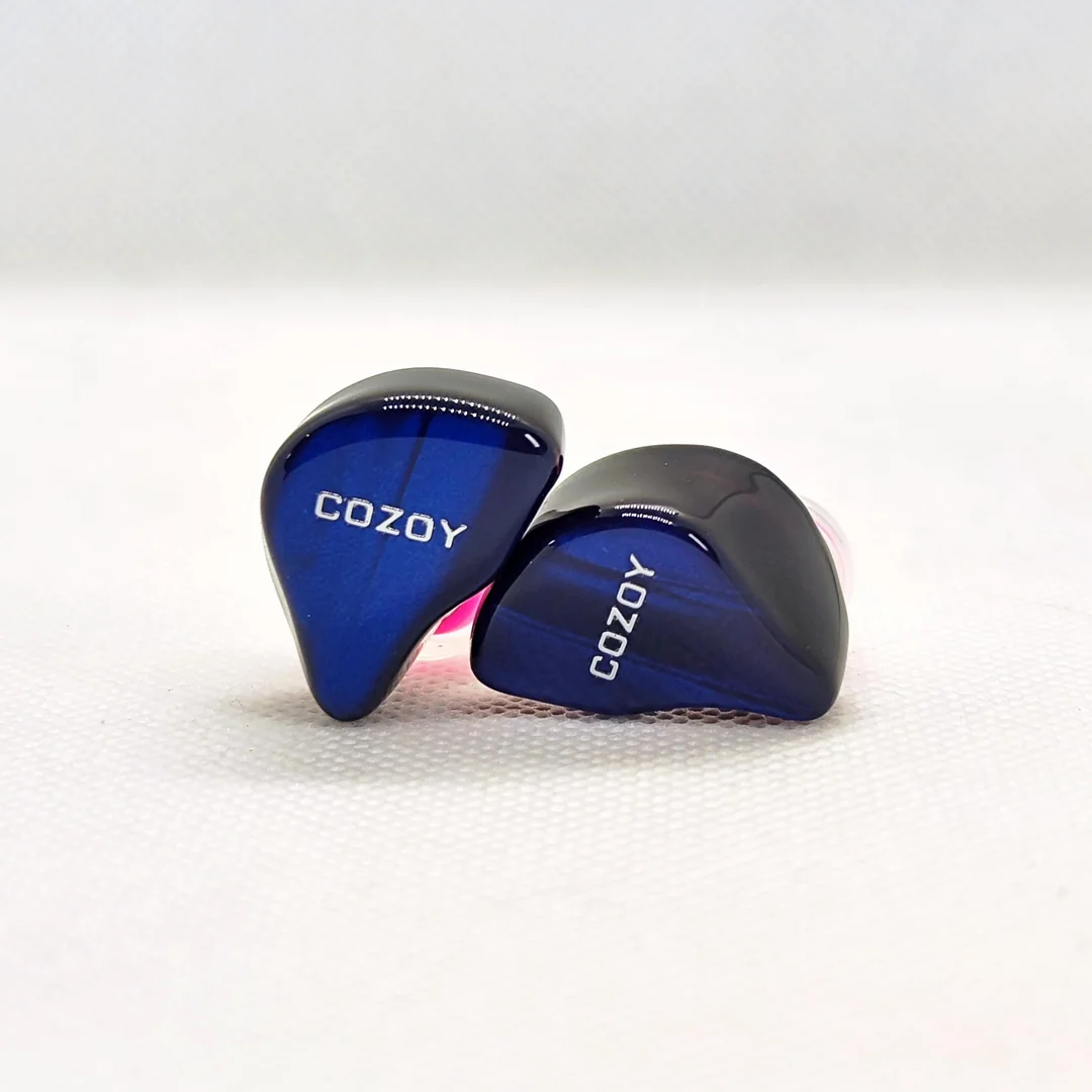 A macro photograph of the two Cozoy D1 IEM shells resting on a white textured background. The image highlights the deep glossy blue faceplates with subtle dark striations, the silver "COZOY" branding, and the ergonomic 3D-printed black resin inner body.