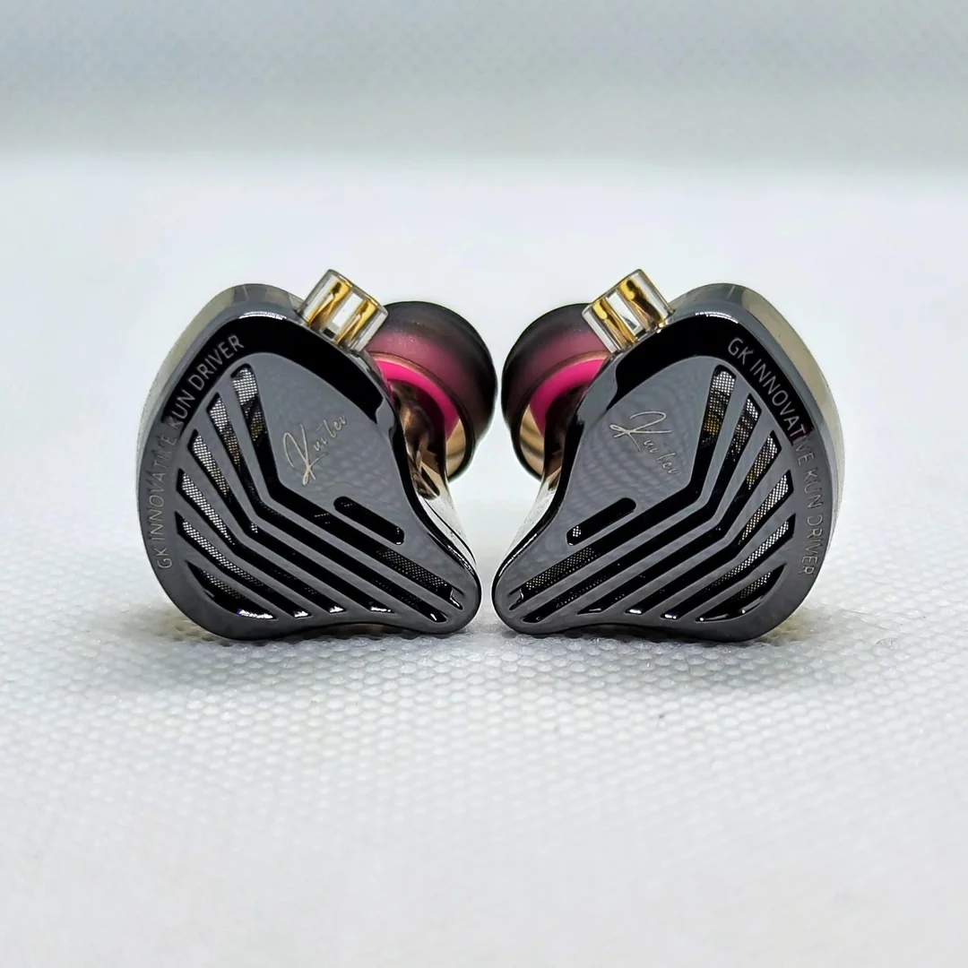 A front-facing macro view of the left and right GK KUNTEN IEM shells side-by-side. The photograph perfectly captures the industrial aesthetic of the dark faceplates with their distinctive cutout vents over acoustic mesh, the white cursive branding, and the protruding transparent QDC-style 2-pin connectors at the top.
